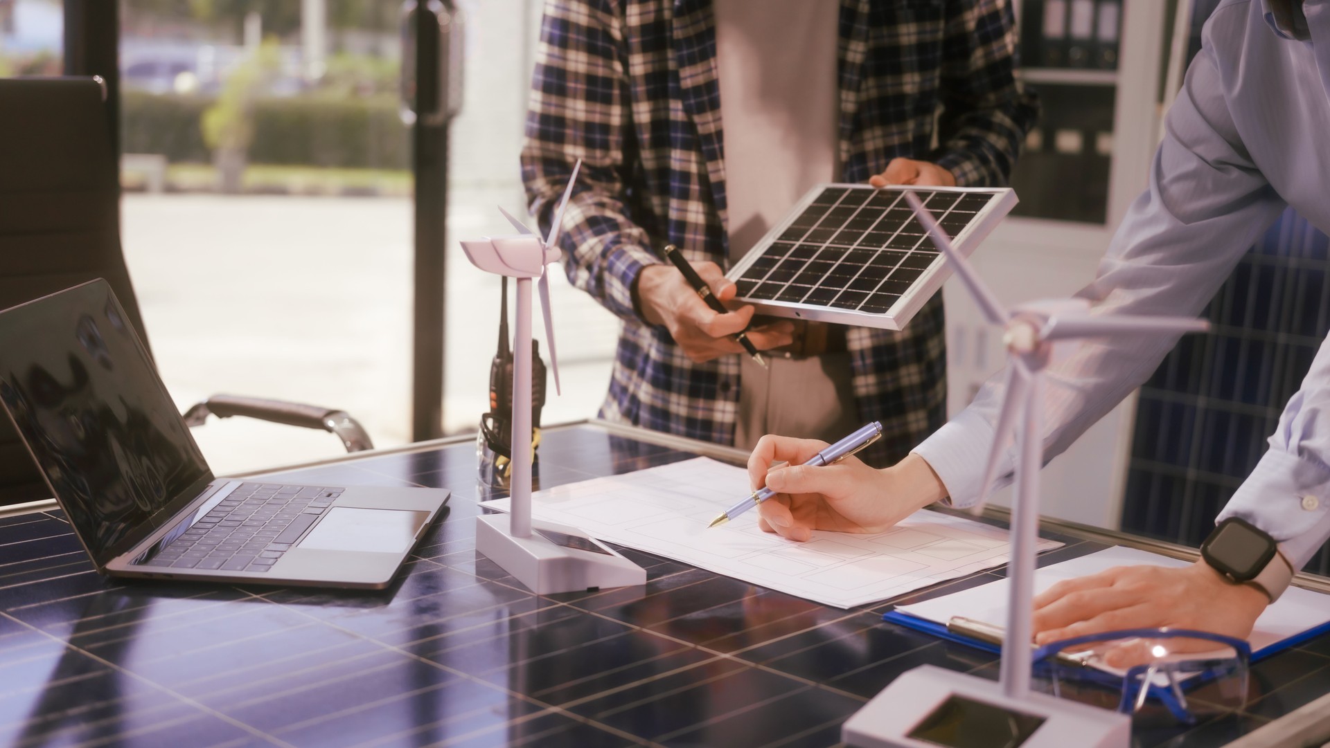 Two Asian businessmen wearing engineer hats table, discussing solar panels and wind turbines. They plan environmental projects, focusing on solar energy solutions and renewable energy installations